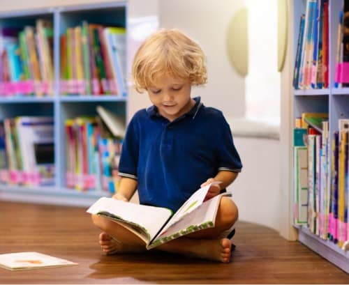 a small child sits on the floor of a library or classroom reading a book