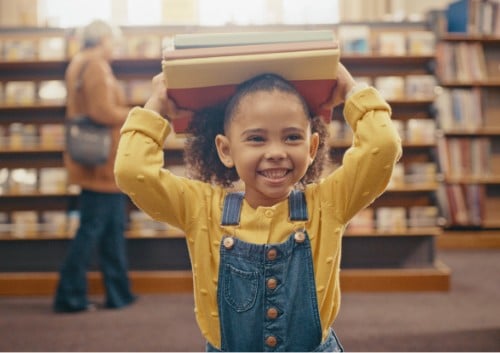 a young girl in a yellow shirt and blue overalls holds a stack of books on her head and smiles in a library setting