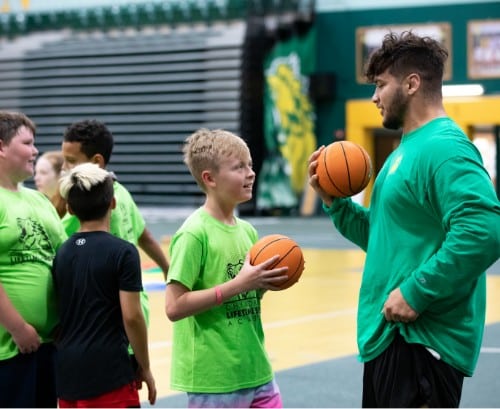 a coach or young adult leader speaks with kids while holding basketballs in a gym