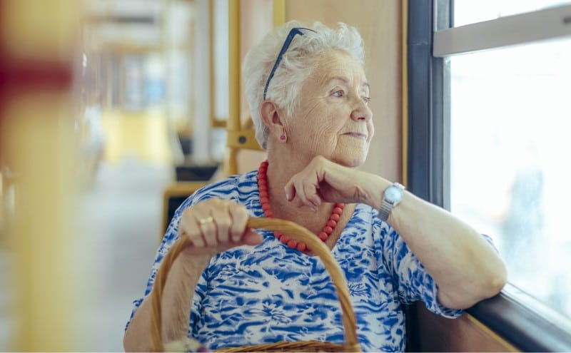 An older woman holding a basket looks out a bus window with her elbow leaned against the sill
