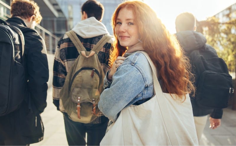 a young female student with long red hair looks back with a smile as three young male students walk in a line just ahead of her