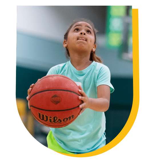 A young girl in action about to shoot a basketball looks up toward the hoop with her tongue out in concentration